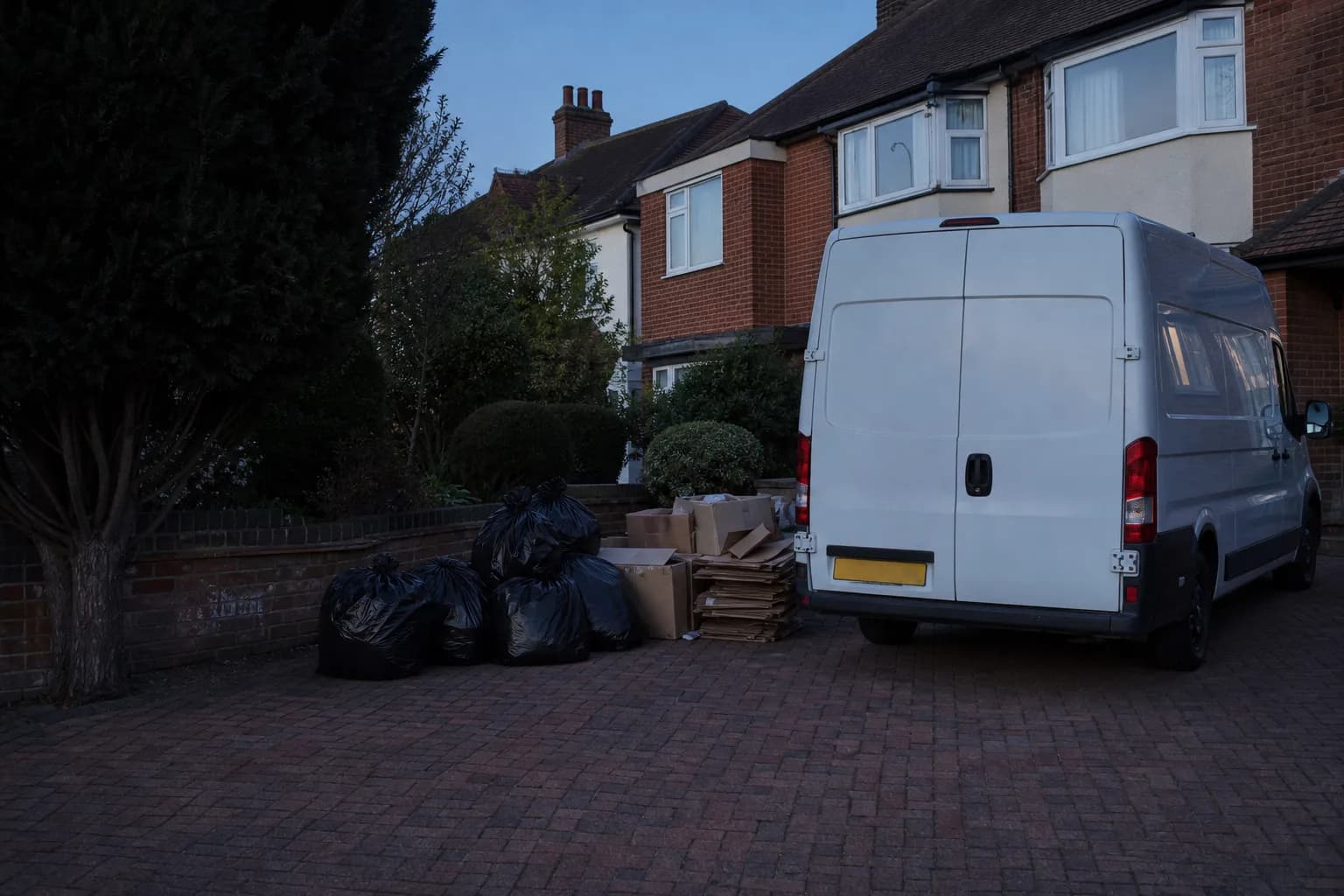 Bagged household waste and flattened cardboard gathered on a suburban driveway beside an unmarked van at dusk
