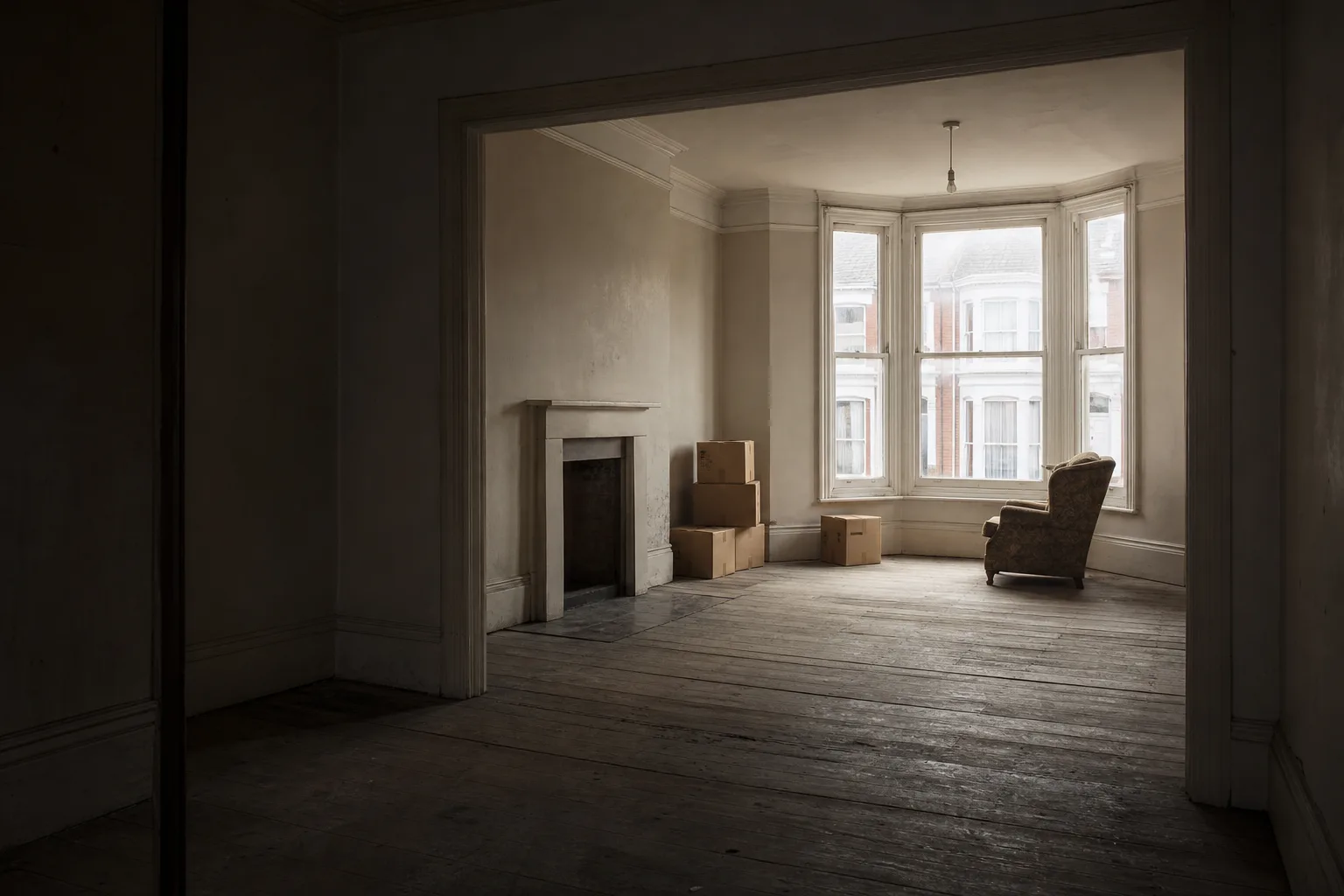A part cleared room in a terraced house with bare floorboards, stacked boxes and light through a bay window